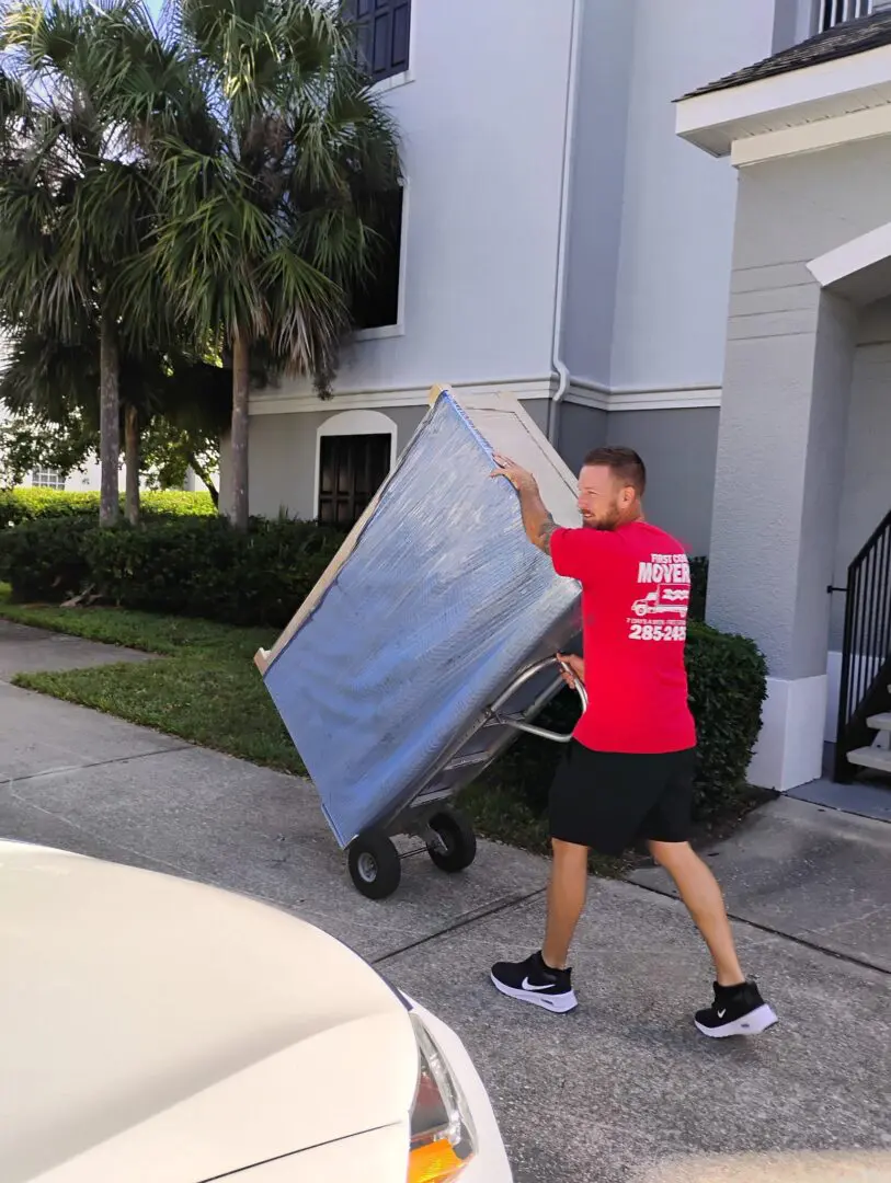 Man in a shirt pushing a trolley full of furniture down a street in, street is lined with trees