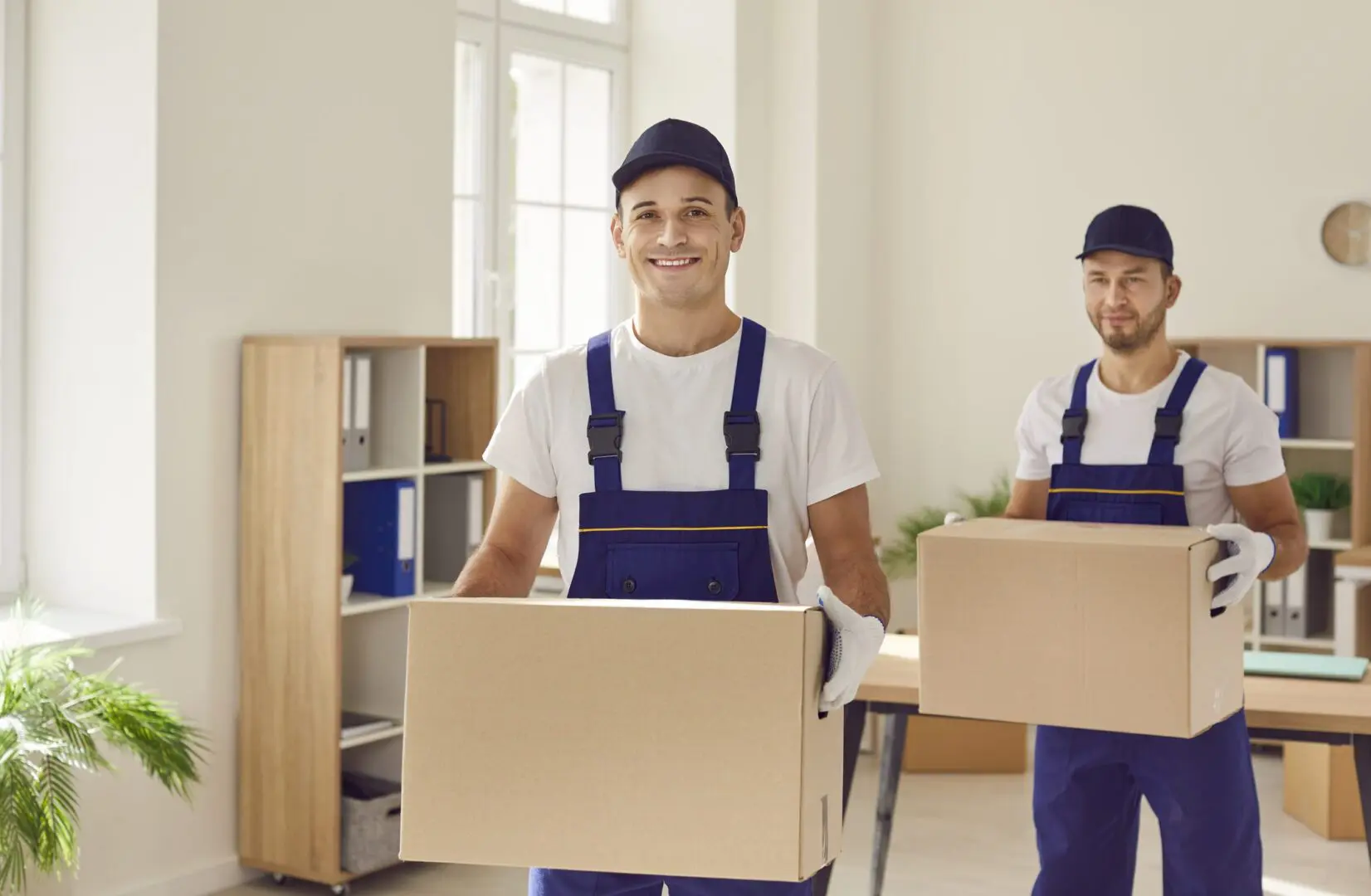 Two movers carrying boxes indoors with smiles.