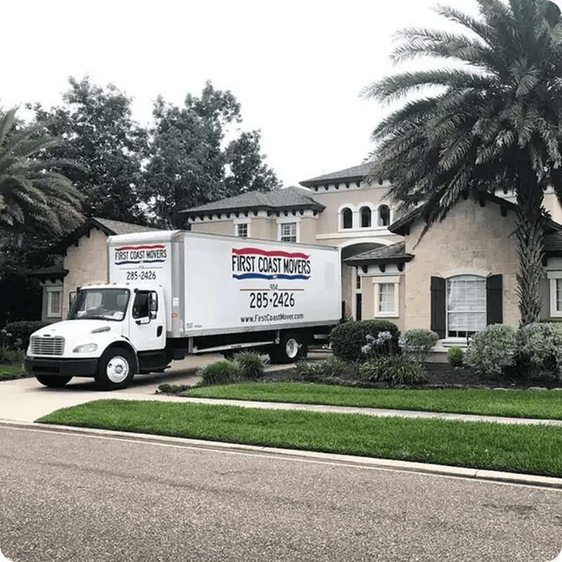 Moving truck parked in front of a house with windows, surrounded by grass, plants, and trees